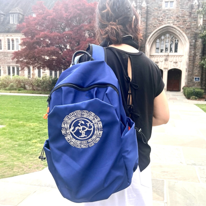 A person viewed from behind wearing a royal blue backpack with a GoldenCrow logo on the front, standing outdoors in front of a stone building and green lawn.