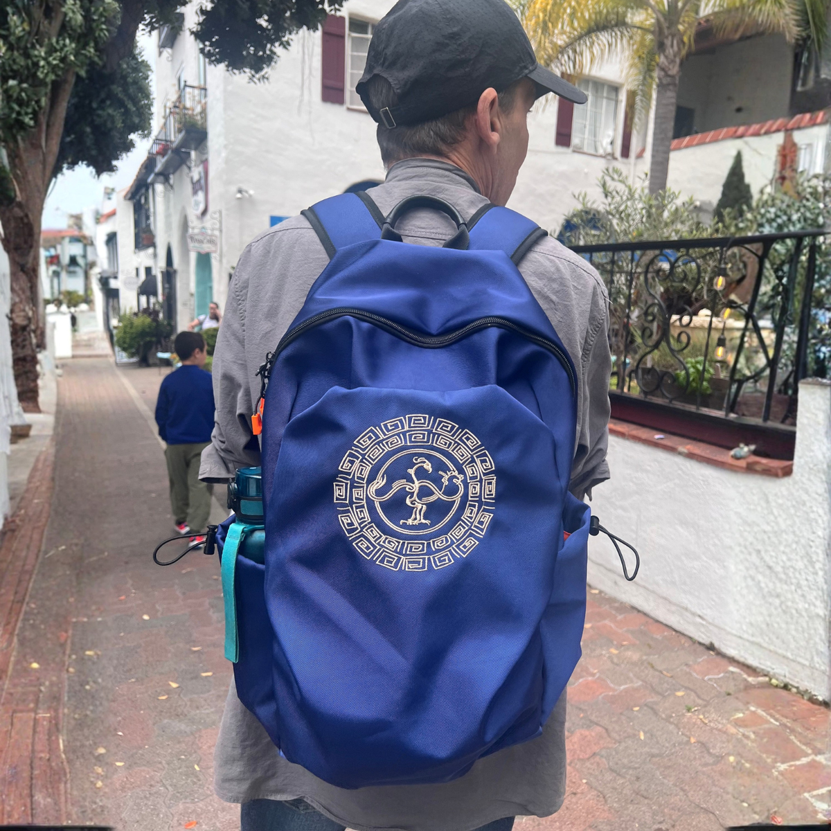 A man viewed from behind wearing a royal blue backpack withGoldenCrow emblem on the back, walking on a brick sidewalk in a residential outdoor setting.