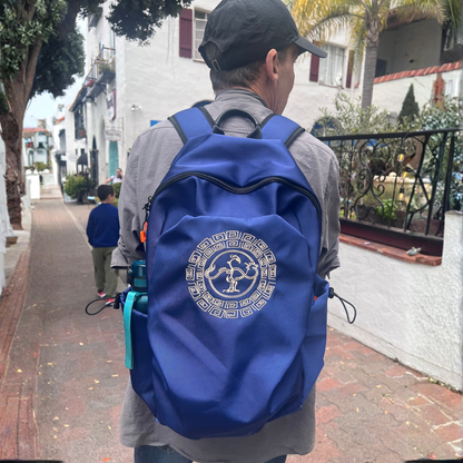 A man viewed from behind wearing a royal blue backpack withGoldenCrow emblem on the back, walking on a brick sidewalk in a residential outdoor setting.