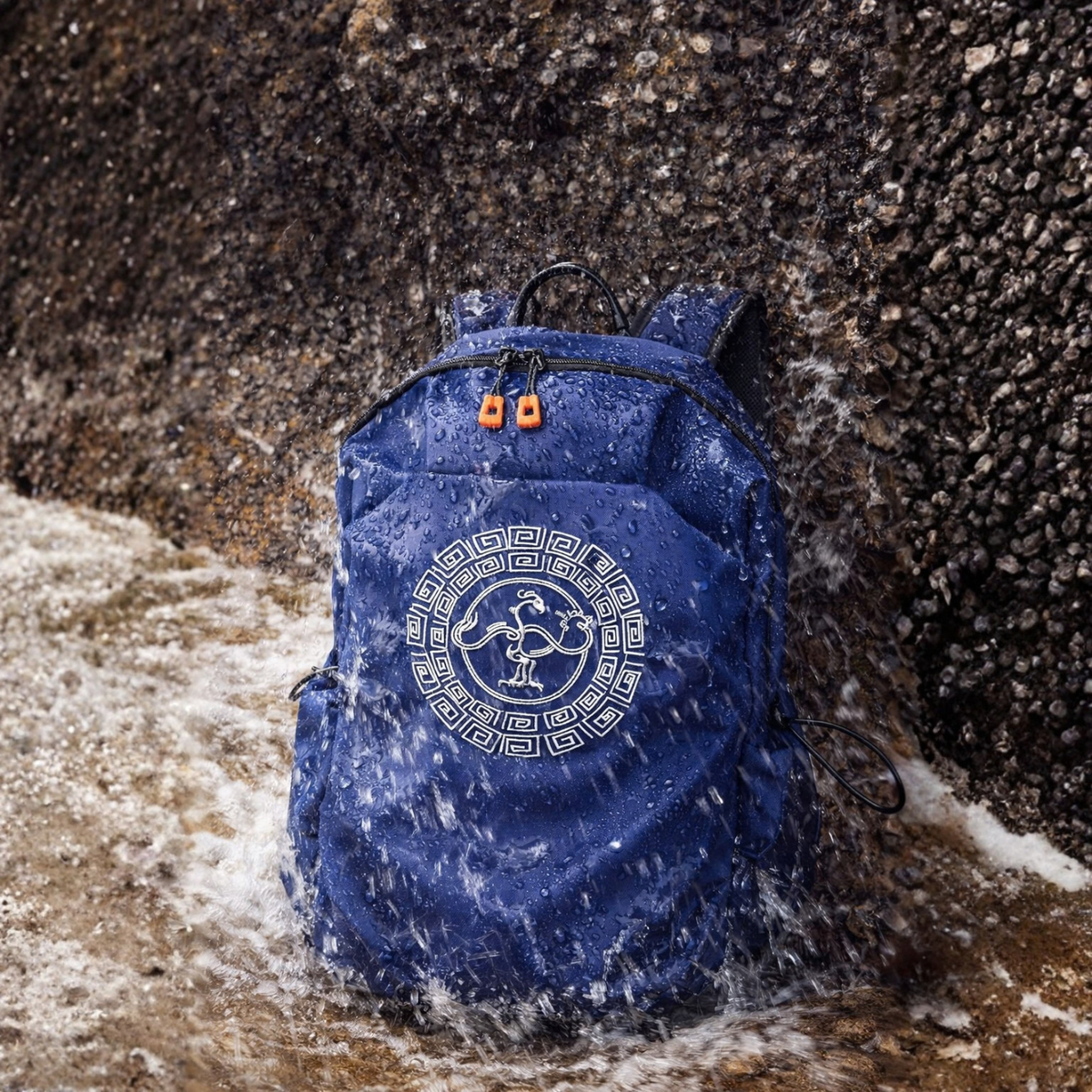 A blue waterproof backpack with a classic GoldenCrow on the front, shown standing in shallow water against a dark rocky shore.