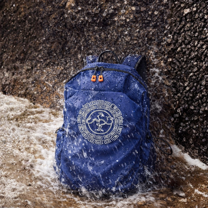 A blue waterproof backpack with a classic GoldenCrow on the front, shown standing in shallow water against a dark rocky shore.