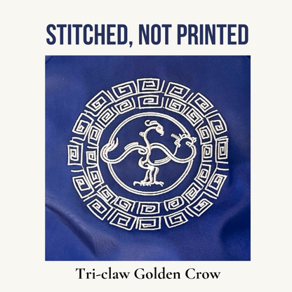 A square product graphic showing a blue fabric item with a golden embroidered circular motif featuring a tree feet crow and geometric border. Large text above reads 'STITCHED, NOT PRINTED' and text below reads 'Tri-claw Golden Crow'.